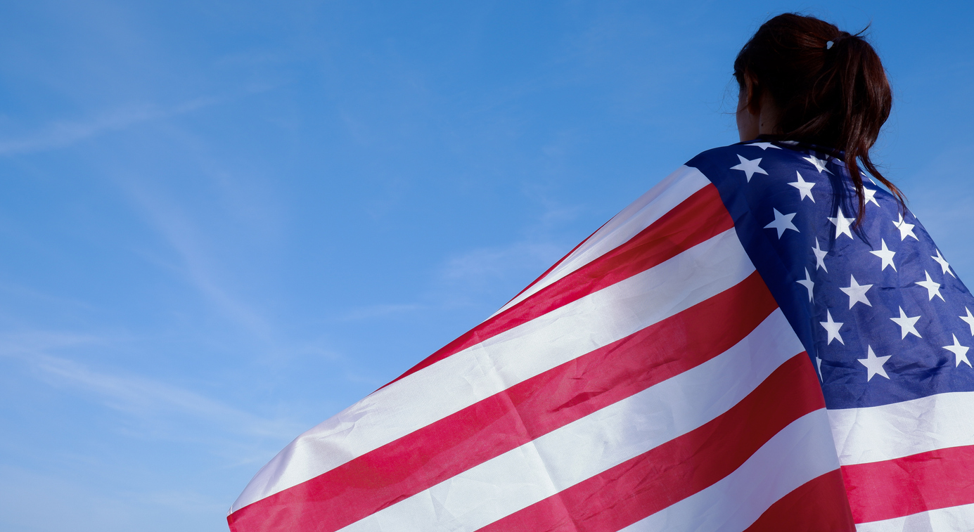 Rear view of a young woman holding an American flag on her shoulders against a blue sky background
